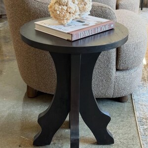 May include: A round black wooden side table with three curved legs. Resting on the tabletop is a hardcover book titled TRAVEL HOME and a textured, off-white, coral-like decorative object. The table features a smooth, circular top surface.