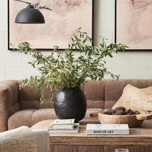 May include: A living room scene featuring a brown sofa, a wooden coffee table, and a black vase filled with greenery. The table holds books and a wooden bowl with pinecones. Artwork hangs on the white brick wall.