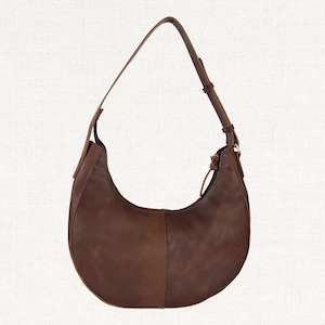 Puede incluir: Un bolso de hombro en forma de media luna de cuero marr&oacute;n con una correa larga y ajustable. El bolso tiene un cierre de cremallera y un dise&ntilde;o simple y minimalista. El cuero tiene un aspecto natural y ligeramente desgastado.