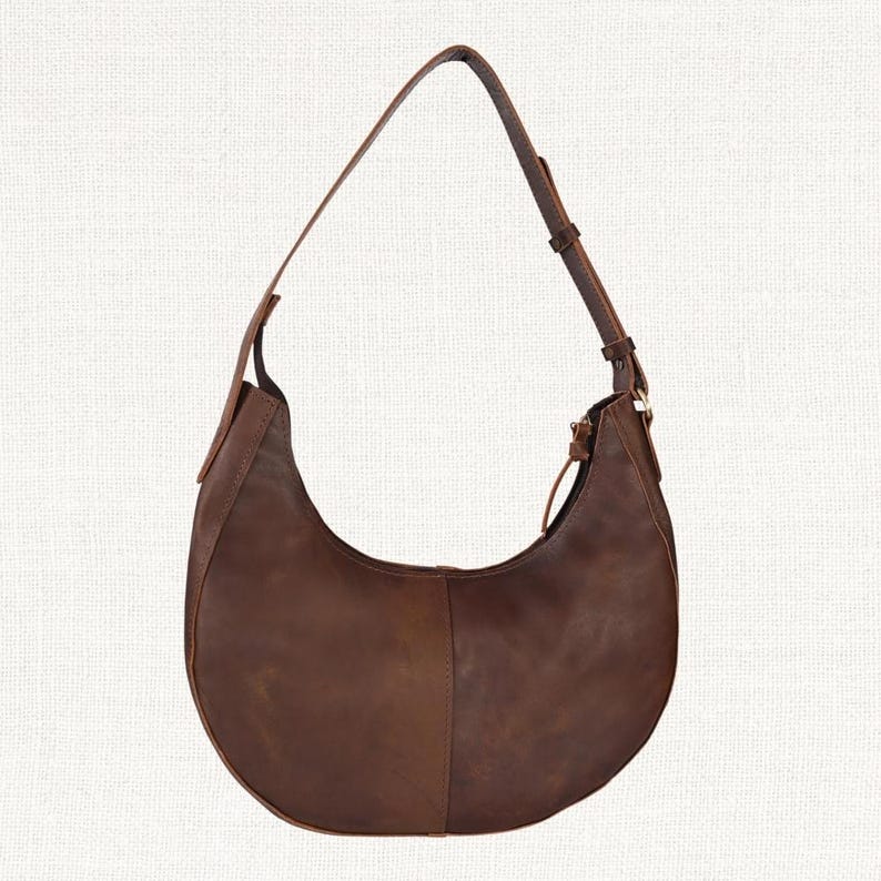 Puede incluir: Un bolso de hombro en forma de media luna de cuero marr&oacute;n con una correa larga y ajustable. El bolso tiene un cierre de cremallera y un dise&ntilde;o simple y minimalista. El cuero tiene un aspecto natural y ligeramente desgastado.