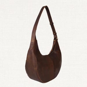 Puede incluir: Un bolso hobo de cuero marr&oacute;n con una sola correa de hombro ajustable. El bolso tiene una forma curva de media luna y un acabado liso, ligeramente desgastado. La correa se sujeta con una hebilla color lat&oacute;n.