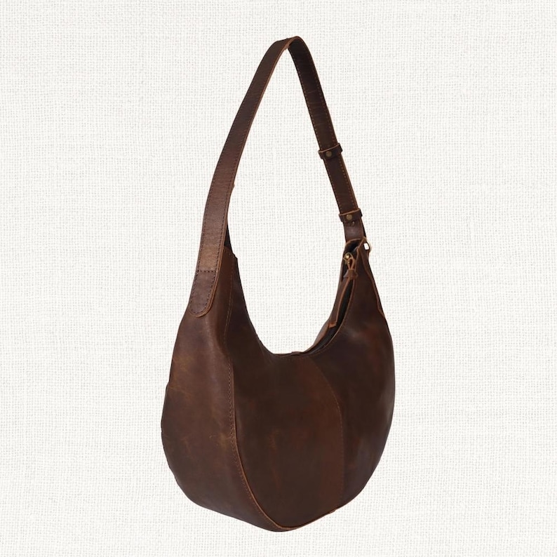 Puede incluir: Un bolso hobo de cuero marr&oacute;n con una sola correa de hombro ajustable. El bolso tiene una forma curva de media luna y un acabado liso, ligeramente desgastado. La correa se sujeta con una hebilla color lat&oacute;n.
