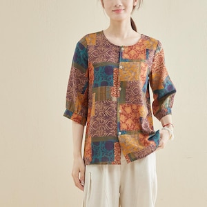 May include: A woman wearing a patchwork print blouse with three-quarter sleeves and button-down front. The blouse features a mix of colors including orange, blue, and brown. She is wearing cream-colored cropped pants and beige flats. The background is a solid light beige.