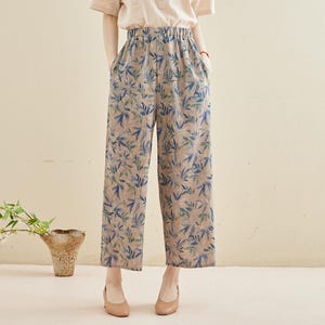 May include: A woman wearing a beige short-sleeved shirt with wooden buttons and blue and green floral print pants. The outfit is paired with beige shoes. The background is a light beige color.
