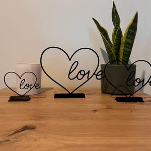 Puede incluir: Tres decoraciones de corazón de metal negro con la palabra "love" en escritura cursiva. Los corazones están sobre una superficie de madera, con una planta en maceta y un recipiente de cerámica a rayas en el fondo.