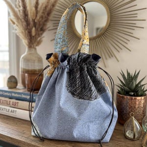 May include: A drawstring tote bag with a light blue speckled fabric body and patterned handles. The bag features a dark gray and black circular design on the front. Brown drawstrings and a dark blue trim complete the design.