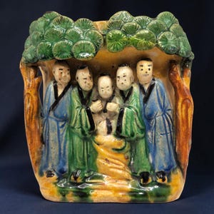May include: A glazed ceramic vessel featuring a relief sculpture of five figures standing beneath a canopy of green pine trees. The figures wear blue and green robes with black trim. The base and tree trunks are rendered in shades of yellow and brown.