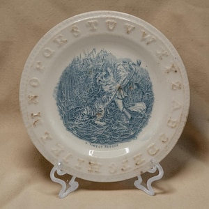 May include: A round white ceramic plate featuring a blue transferware illustration of a person and a tiger in tall grass. The text A TIMELY RESCUE is printed below the scene. The rim is embossed with the alphabet letters from A through Z.