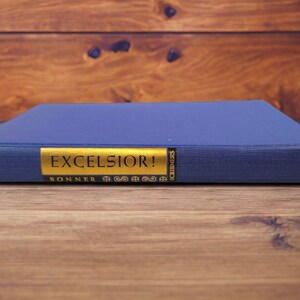 Naked book spine – Blue cloth-bound spine with bright gold title strip reading “Excelsior!” and “Scribners.”
