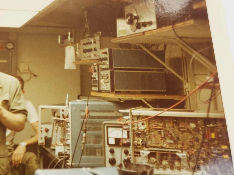 May include: A vintage photo of a room with electronic equipment, including a large, multi-layered device with knobs and dials, and a smaller device with a digital display. The room appears to be a laboratory or workshop.