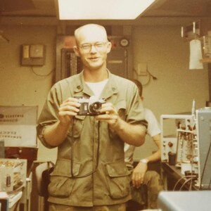 May include: A man in a green military uniform holds a camera in front of him. He is smiling and looking at the camera. The background is a room with various electronic equipment.