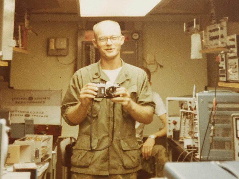 May include: A man in a green military uniform holds a camera in front of him. He is smiling and looking at the camera. The background is a room with various electronic equipment.