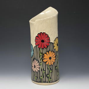 Puede incluir: Una taza de cerámica blanca con un asa negra y el texto "But First, Coffee" en negrita negra. La taza se encuentra sobre un fondo borroso, sugiriendo un ambiente acogedor.