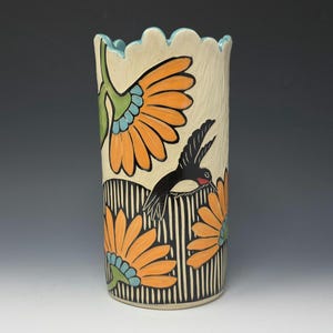 Puede incluir: Un jarrón de cerámica alto con fondo crema, adornado con flores naranjas, un pájaro negro y rojo y patrones de rayas blancas y negras. El borde es festoneado y pintado de azul claro.