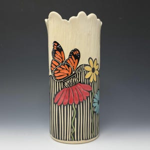 Puede incluir: Un jarrón de cerámica alto, de color crema, con un borde festoneado. El jarrón presenta un diseño pintado de una mariposa monarca, flores rojas y amarillas, y un fondo a rayas blancas y negras. El jarrón mide unos 25 cm de alto.