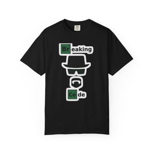 Może przedstawiać: Czarna koszulka z białym i zielonym wzorem graficznym. Wzór przedstawia słowa "Breaking Code" z ilustracją kapelusza, okularów i brody. Koszulka ma krótki rękaw i okrągły dekolt.