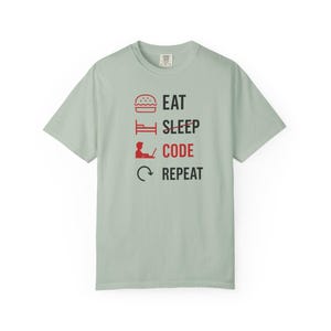 May include: A pale green t-shirt featuring the text "EAT SLEEP CODE REPEAT" in black and red. The design includes a burger, bed, person with a laptop, and a circular arrow. The shirt is made of a soft material.
