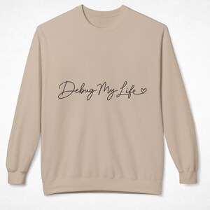 May include: A beige long-sleeve crewneck sweatshirt featuring the black cursive text Debug My Life with a small heart symbol at the end. The garment has a ribbed collar, cuffs, and hem, providing a clean and simple look for casual wear.