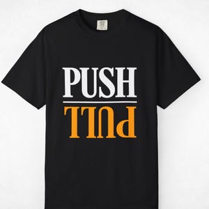May include: A black short-sleeved t-shirt featuring the word PUSH in white capital letters above a horizontal line, with the word PULL printed in orange capital letters below the line in an inverted, upside-down orientation.