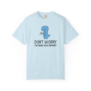 Könnte beinhalten: Hellblaues T-Shirt mit einem Cartoon-Dinosaurier, der eine Kaffeetasse hält. Der Text "DON'T WORRY I'M FROM TECH SUPPORT" ist darunter gedruckt. Das T-Shirt besteht aus einem weichen Material.