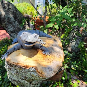 May include: A gray and white lizard figurine with articulated limbs rests on a tree stump. The lizard has a smiling expression and is surrounded by green foliage and a tree trunk. The scene is set outdoors in natural light.