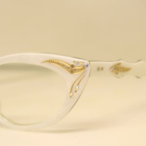 May include: White cat-eye glasses with gold and rhinestone embellishments. The glasses have a delicate, feminine design.