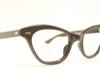 May include: Black cat-eye eyeglasses with a classic retro design. The frames are a glossy black, featuring a subtle decorative detail on the temples. The lenses are clear, and the overall style is reminiscent of vintage eyewear.