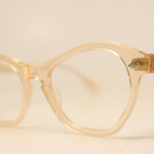Puede incluir: Gafas de color melocotón pálido con forma de ojo de gato. Las monturas son de plástico translúcido con pequeños detalles metálicos decorativos en las patillas. Las lentes son transparentes y el diseño general es de inspiración vintage.