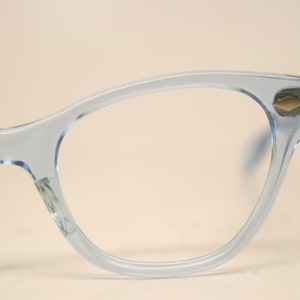 Puede incluir: Primer plano de gafas azul claro con forma de ojo de gato. Las monturas son de color azul claro translúcido. Las patillas tienen un pequeño acento dorado. Las lentes son transparentes. Las gafas están sobre un fondo neutro.