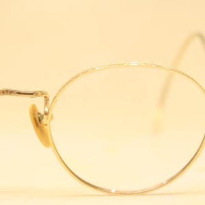 May include: Close-up of vintage gold-tone eyeglasses with round lenses. The frames have a delicate, etched design along the top and temples. The nose pads are a light tan color, and the earpieces curve gently.
