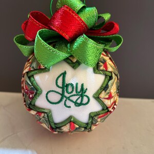 May include: A handcrafted Christmas ornament with a festive design. The ornament features a white starburst shape with the word "Joy" embroidered in green. It is adorned with red and green ribbons and a green hanging string. The ornament is approximately 3 inches in diameter.