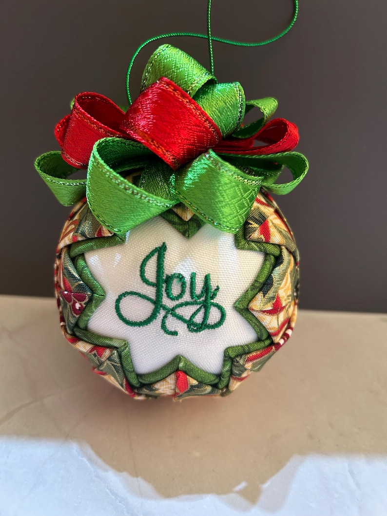 May include: A handcrafted Christmas ornament with a festive design. The ornament features a white starburst shape with the word "Joy" embroidered in green. It is adorned with red and green ribbons and a green hanging string. The ornament is approximately 3 inches in diameter.