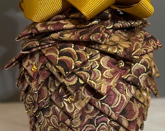 May include: A decorative ornament shaped like a pinecone, featuring a quilted design in burgundy, gold, and brown tones. The ornament is topped with a large gold ribbon bow and has a gold hanging loop. Small gold beads are scattered on the surface.