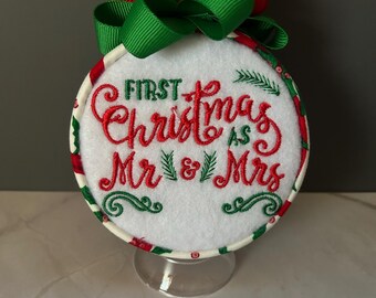 May include: Embroidered Christmas ornament with the text "First Christmas as Mr & Mrs" in red script. The ornament has a white background, a red and green border, and a red and green bow at the top. A clear stand supports the ornament.