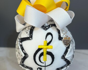 May include: A fabric ornament with a musical theme. The white ball features a yellow cross and treble clef, with a black and white musical note pattern. It is topped with a yellow and white bow and a silver charm.