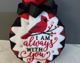 May include: A round, fabric ornament with a red cardinal bird design and the text "I AM always WITH you". The ornament has a white background, red and black accents, and a red and black bow at the top. Rhinestones are placed around the design.
