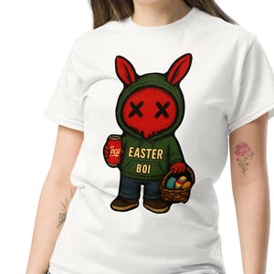 Puede incluir: Camiseta blanca con un personaje de conejo de Pascua de dibujos animados. El conejo tiene ojos y orejas rojas, una sudadera con capucha verde y sostiene una lata de refresco y una cesta de huevos. El texto "EASTER BOI" está en la sudadera.