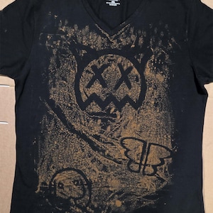 Könnte beinhalten: Ein schwarzes T-Shirt mit V-Ausschnitt und einem beigefarbenen Spritzlack-Effekt auf der Vorderseite. Die Grafik zeigt ein stilisiertes katzenartiges Gesicht mit X-förmigen Augen, eine kleine kreisförmige Figur und einen Schmetterlingsumriss. Das Innenetikett zeigt Jack Parker.