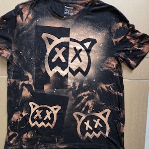 Może przedstawiać: Czarny t-shirt z krótkim rękawem z efektem wybielenia, pomarańczowo-brązowymi rozpryskami oraz trzema grafikami kocich twarzy z oczami w kształcie litery X. Koszulka posiada okrągły dekolt i nadrukowaną metkę marki Parkside na wewnętrznym kołnierzu.