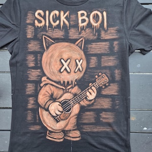 Puede incluir: Una camiseta negra de manga corta con un estampado gráfico de un personaje felino con sudadera con capucha tocando la guitarra. El personaje tiene ojos en forma de X sobre un fondo de pared de ladrillo. El texto SICK BOI está impreso arriba.