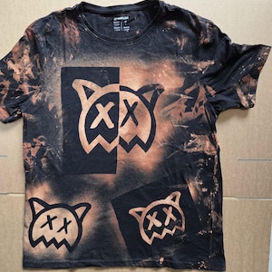 Pode incluir: Uma t-shirt preta de manga curta com um efeito de lixívia e salpicos de cor laranja acastanhada. A peça apresenta três ícones gráficos de uma cabeça de gato estilizada com olhos em forma de X, impressos a preto nas zonas aclaradas.