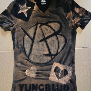 Op de afbeelding: Een zwart T-shirt met V-hals en een gebleekt effect, voorzien van een groot cirkelvormig logo, een ster, een hart en een gebroken hart. De tekst JACK PARKER staat in de binnenkraag en het woord YUNGBLUD is onderaan in het zwart gedrukt.