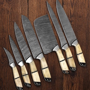 Può includere: Un set di sette coltelli da cucina con lame in acciaio decorate e manici color crema. I coltelli variano per dimensioni e forma, tra cui un coltello da macellaio e diversi coltelli da chef. I manici hanno dettagli neri, su uno sfondo di legno scuro.