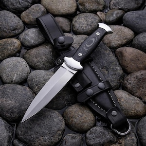 May include: A fixed-blade knife with a double-edged silver steel blade, a polished silver bolster, and a black wood-grain handle secured with two silver rivets. The knife rests on a black leather sheath featuring silver snaps and white stitching.