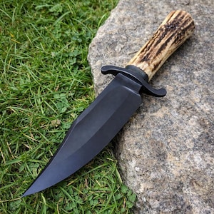 Può includere: Un coltello a lama nera con un manico in corno strutturato. Il coltello ha una lama larga e affilata e una guardia curva. Il manico è di colore marrone chiaro con accenti marrone scuro. Il coltello è appoggiato su una pietra grigia con erba verde sullo sfondo.