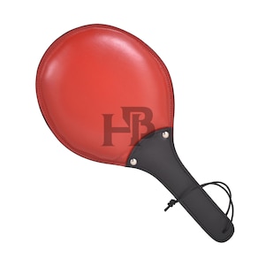 May include: A red leather paddle with a black handle and trim. The paddle has a rounded shape and is stitched around the edges. The handle has a loop for hanging. The letters "HB" are visible on the paddle.