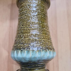 May include: A ceramic vase featuring a textured, olive-green upper body with horizontal ridges and a smooth, flared base glazed in a contrasting light blue. The cylindrical vessel has a wide, rounded rim and a distinct, tiered pedestal foot.