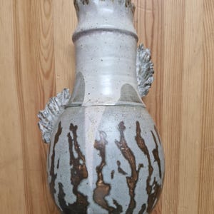 May include: A ceramic vase with a light grey speckled glaze and a bulbous body featuring vertical, dark brown, branch-like markings. The vase has a flared, ruffled rim and two decorative, textured, leaf-shaped protrusions on the sides.
