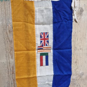 Peut inclure: Un drapeau vintage avec des rayures verticales dorées, blanches et bleues. La bande blanche présente un emblème détaillé avec un Union Jack et d'autres rayures colorées. Le drapeau est en tissu et a un aspect usé.