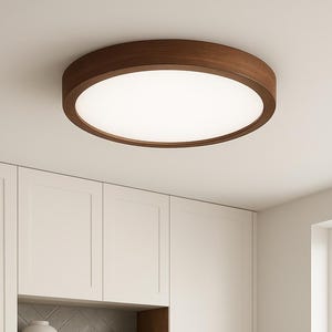 May include: A modern, circular flush-mount ceiling light with a dark wood trim and a bright, white light panel. The light is installed in a kitchen with white cabinets, a gray backsplash, and a dark sink.